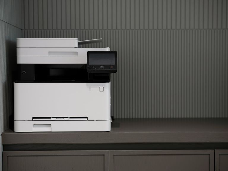 A white office copier on a grey cabinet against a ribbed grey wall.