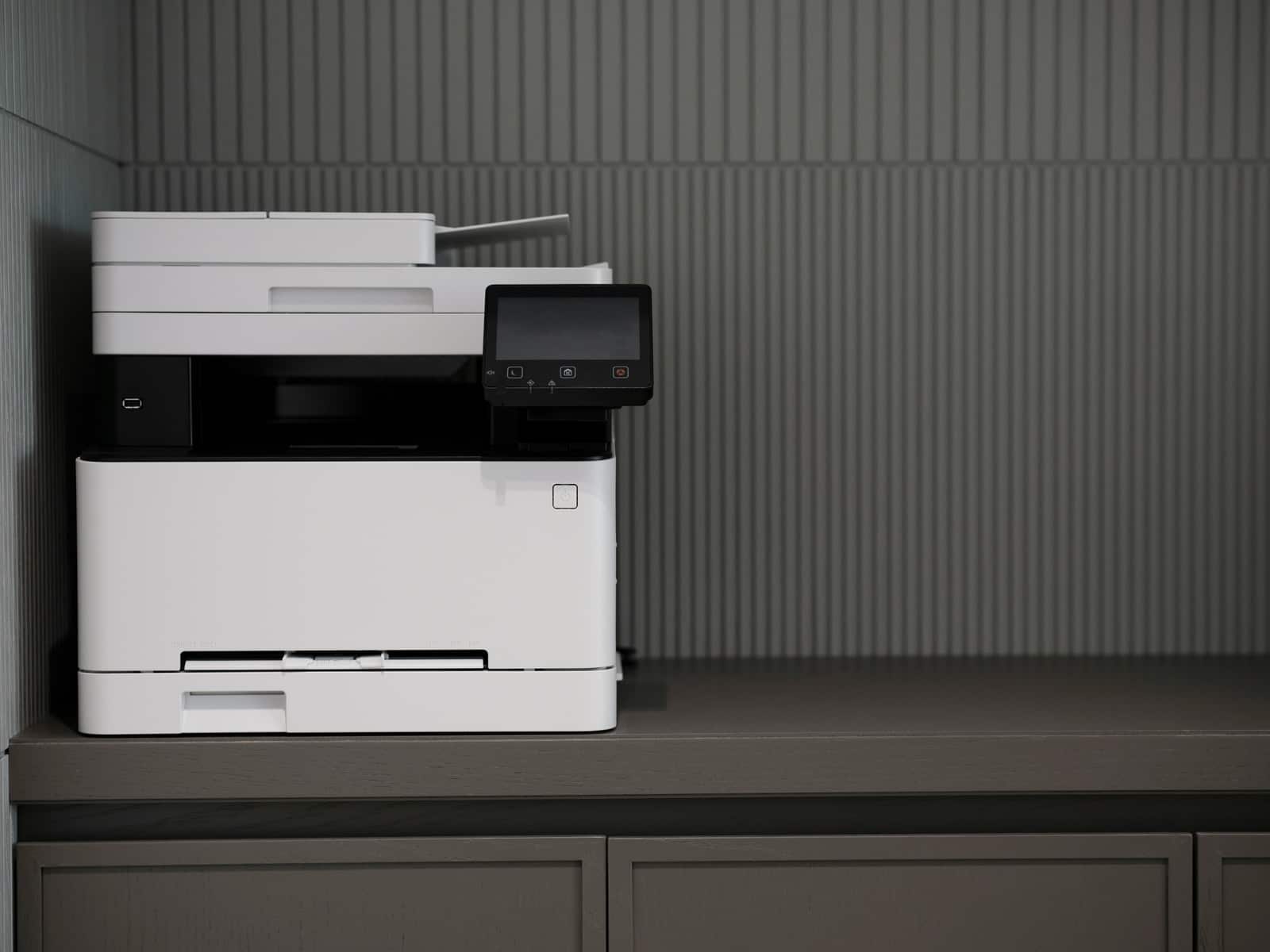 A white office copier on a grey cabinet against a ribbed grey wall.
