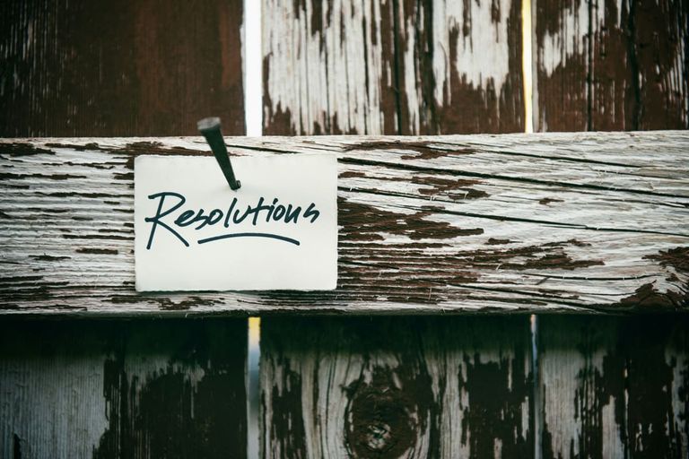 A white card with the handwritten word Resolutions nailed to a weathered wooden fence rail with peeling white paint.