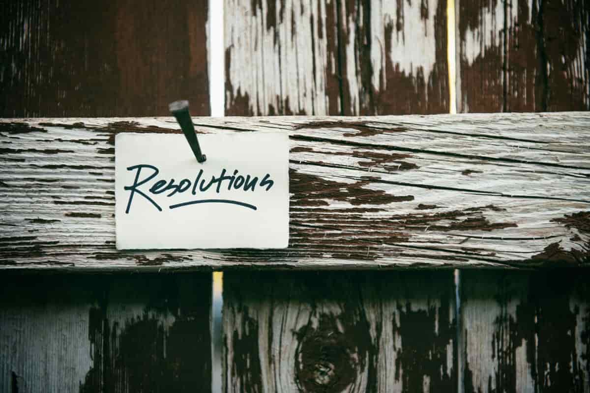 A white card with the handwritten word Resolutions nailed to a weathered wooden fence rail with peeling white paint.