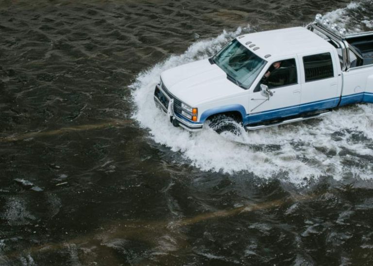 A white pickup truck driving through deep floodwater, throwing up a wake of spray.