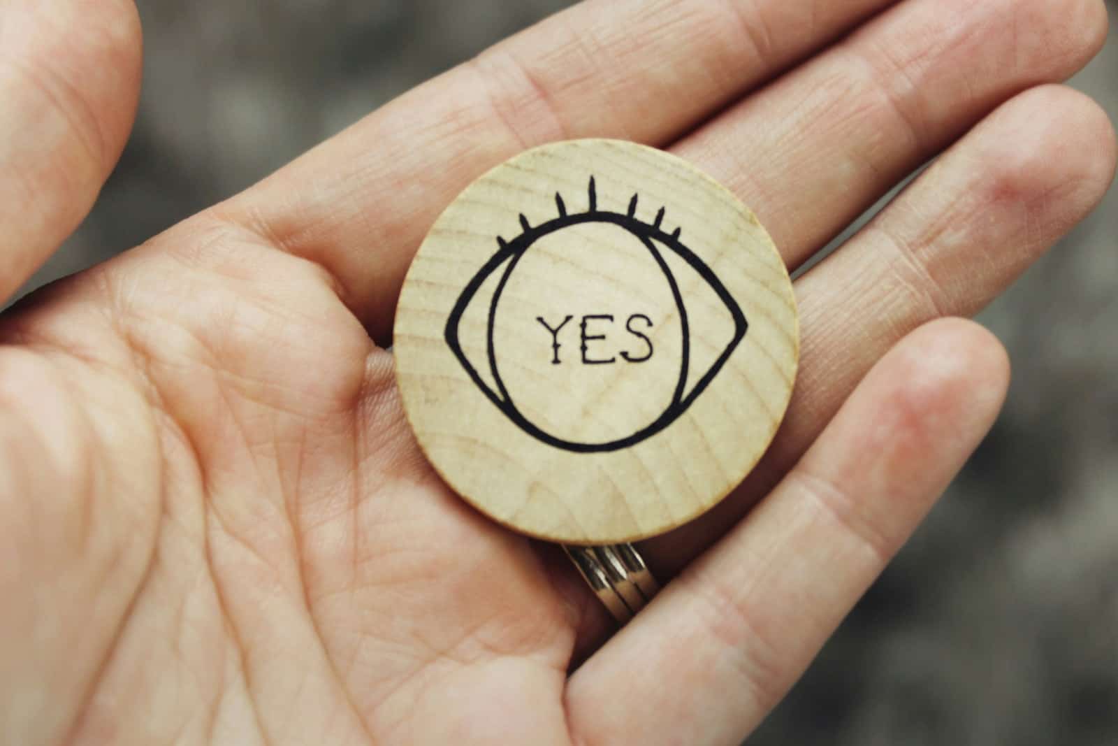 A hand holding a small round wooden token printed with an open eye and the word YES.