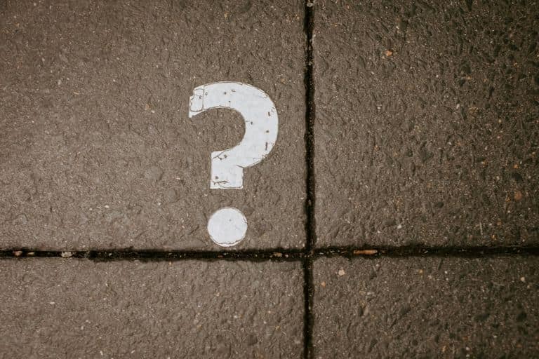 A white question mark painted on a dark paved sidewalk, split by two grout lines.